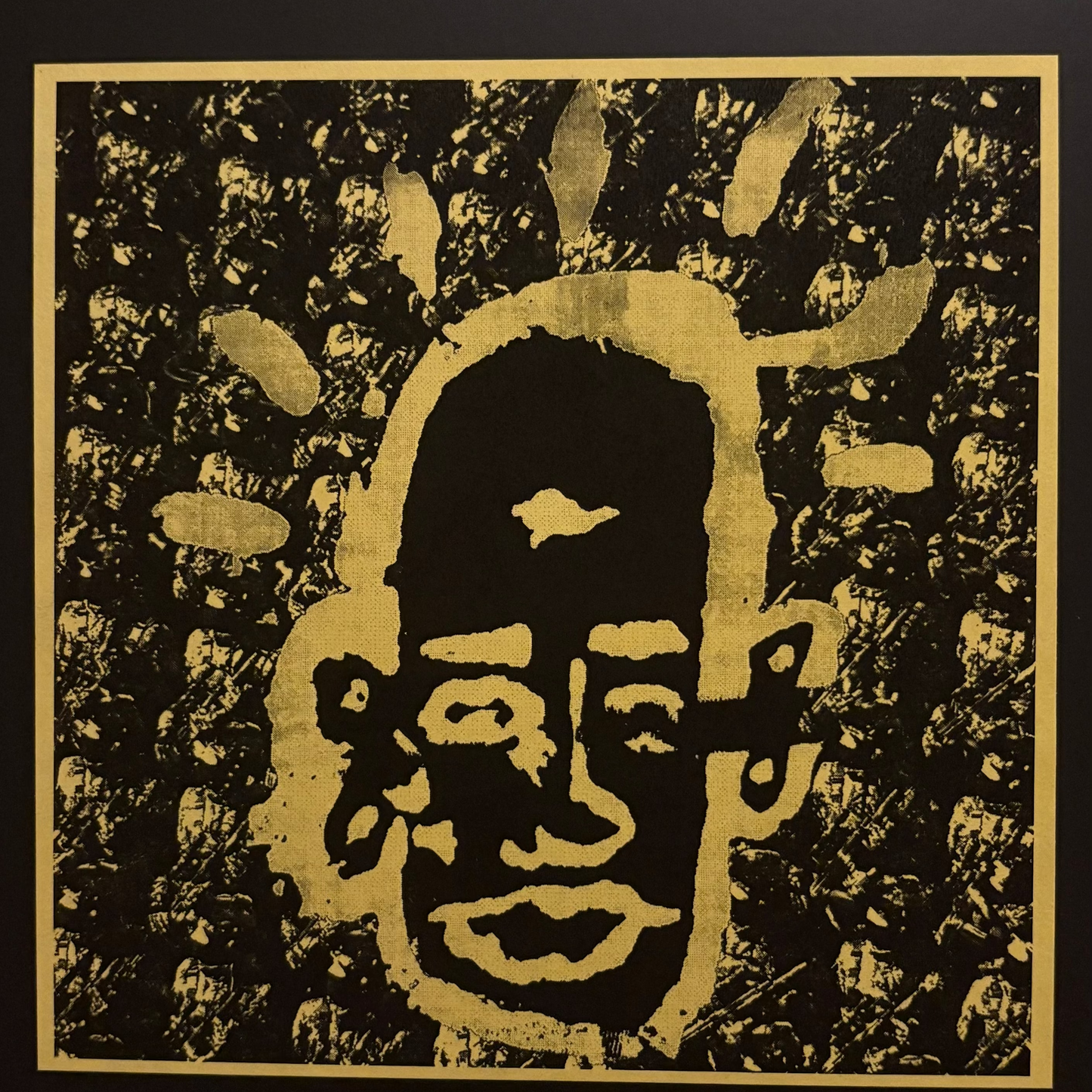 A square black and gold art print featuring a stylized face with radiating gold shapes on a textured black background, bordered by a gold frame against a dark backdrop.